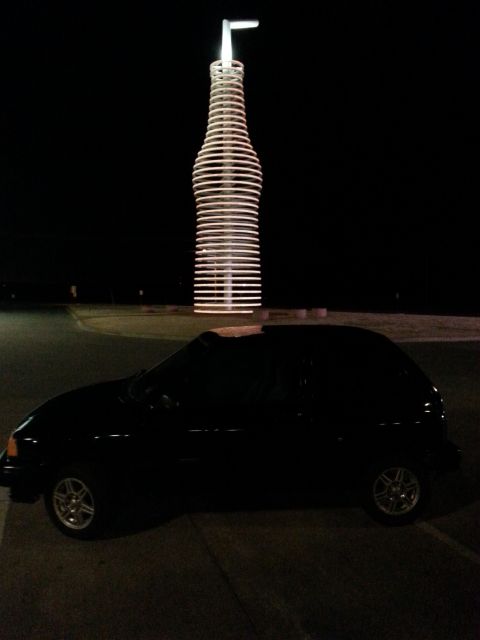 1991 Ford Festiva - A black hatchback is parked at night in front of a large, illuminated bottle-shaped sculpture.