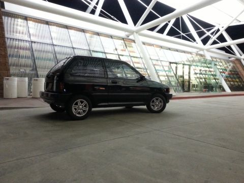 1991 Ford Festiva - A black 1991 Ford Festiva is seen from the side in front of a building with a distinctive white, angular roof structure.