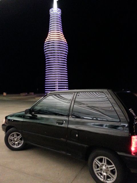 1991 Ford Festiva - A black hatchback is parked at night with a large, illuminated soda bottle sculpture in the background.