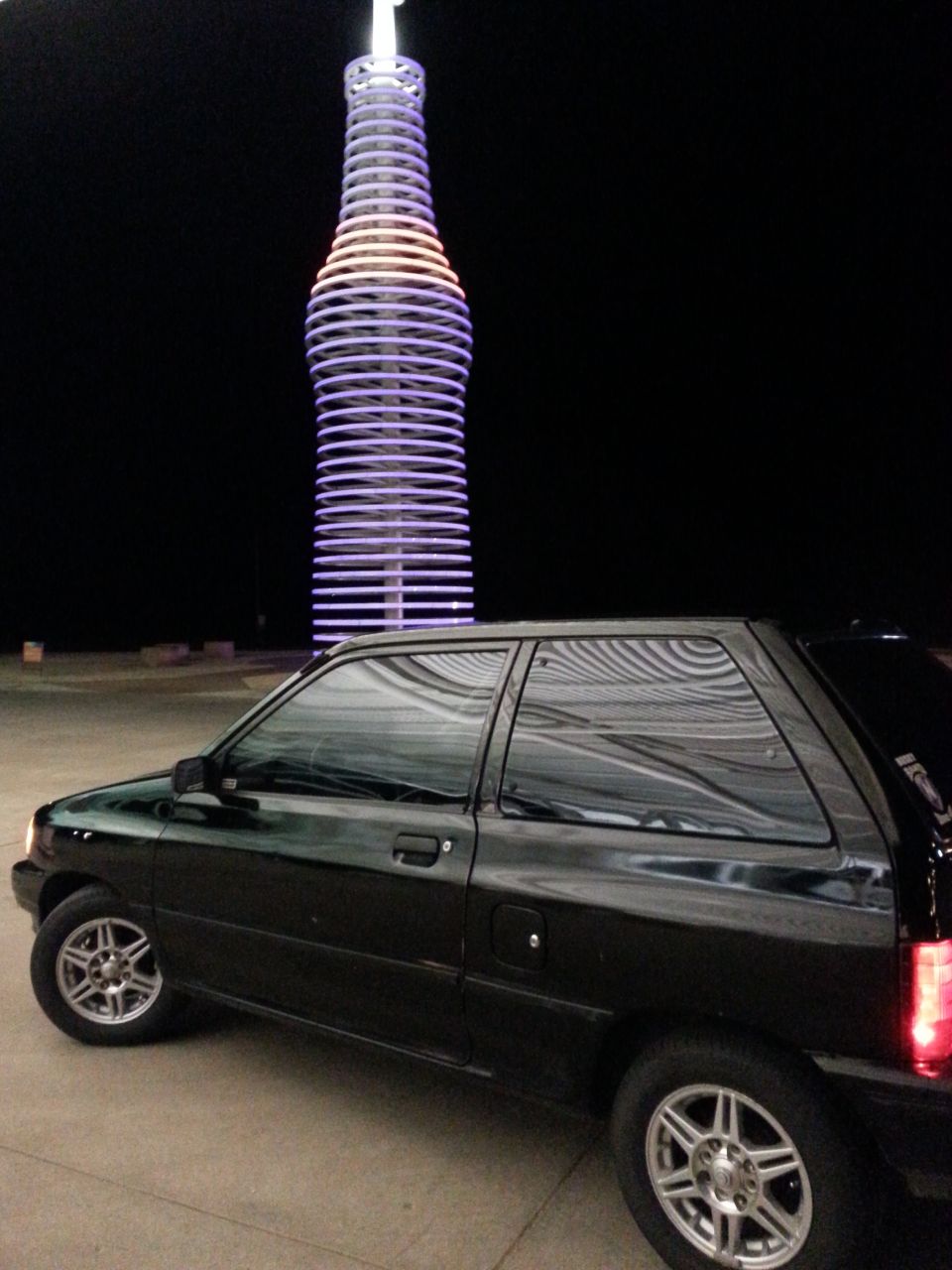 black hatchback parked at night near a giant illuminated soda bottle sculpture