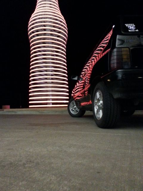 1991 Ford Festiva - A black hatchback with a distinctive zebra stripe decal is parked near a giant illuminated bottle sculpture at night.