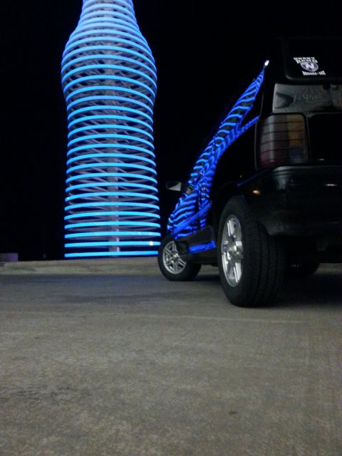 1991 Ford Festiva - A black car with custom blue neon lighting is parked near a large, illuminated bottle structure at night.