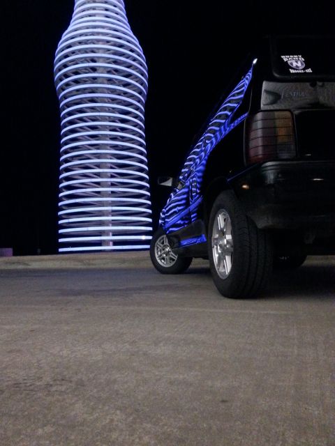 1991 Ford Festiva - A car with custom neon lighting is parked at night next to a large, glowing bottle-shaped sculpture.