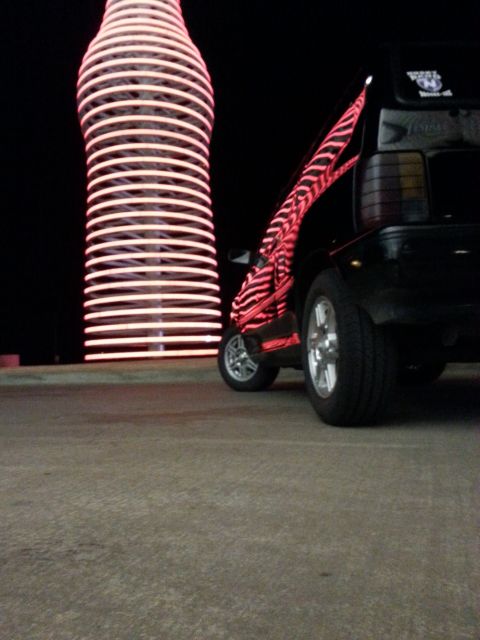 1991 Ford Festiva - A car with a distinctive red zebra stripe decal is parked at night near a large, glowing bottle-shaped landmark.