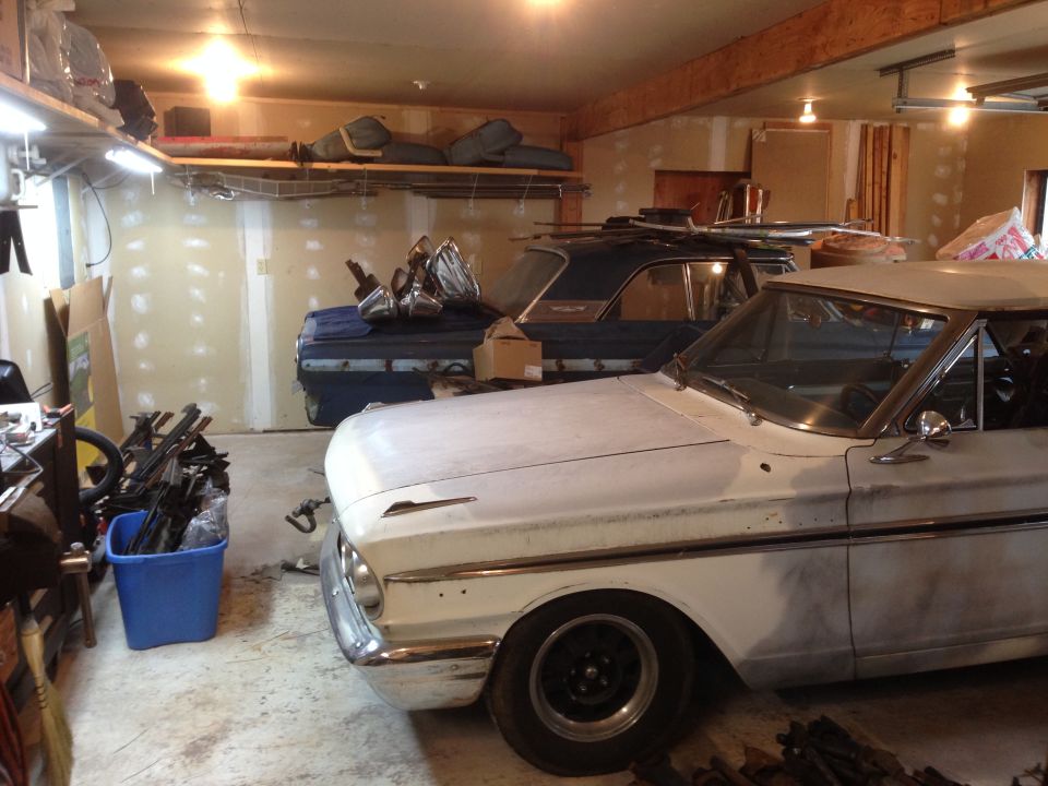 Front of a white 1964 Ford Fairlane, primer finish, next to a dark blue car in a garage