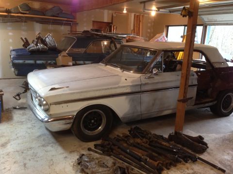 1964 Ford Fairlane - A white 1964 Ford Fairlane convertible sits in a garage next to another vehicle, surrounded by automotive parts.
