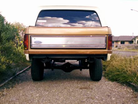 1982 Ford Bronco - Rear view of a gold and white Ford Bronco, showing its differential and taillights on a gravel road.