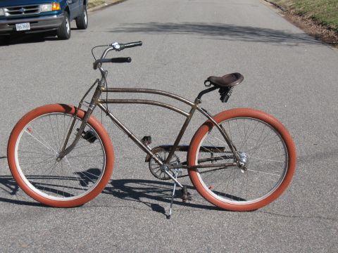 Rusty cruiser bicycle with orange tires parked on asphalt road, blue pickup truck in background