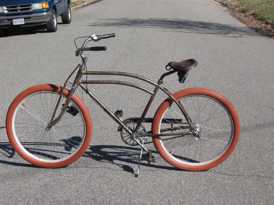 Rusty cruiser bicycle with orange tires parked on asphalt road, blue pickup truck in background