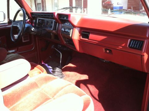1986 Ford Bronco - The interior of a vehicle shows a red dashboard, seats, and a manual transmission shifter.