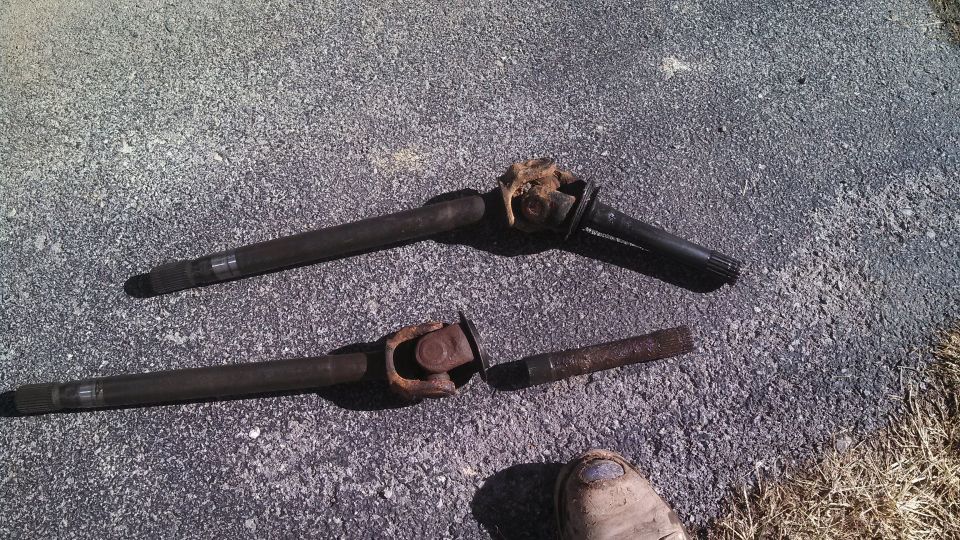 Two rusty driveshafts and a broken piece of driveshaft lying on asphalt, with a boot in the lower right corner