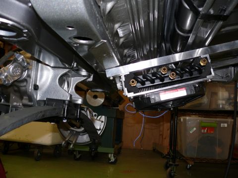 Underside view of vehicle suspension and exhaust system, showing a metal bracket with coiled tubes and a fan