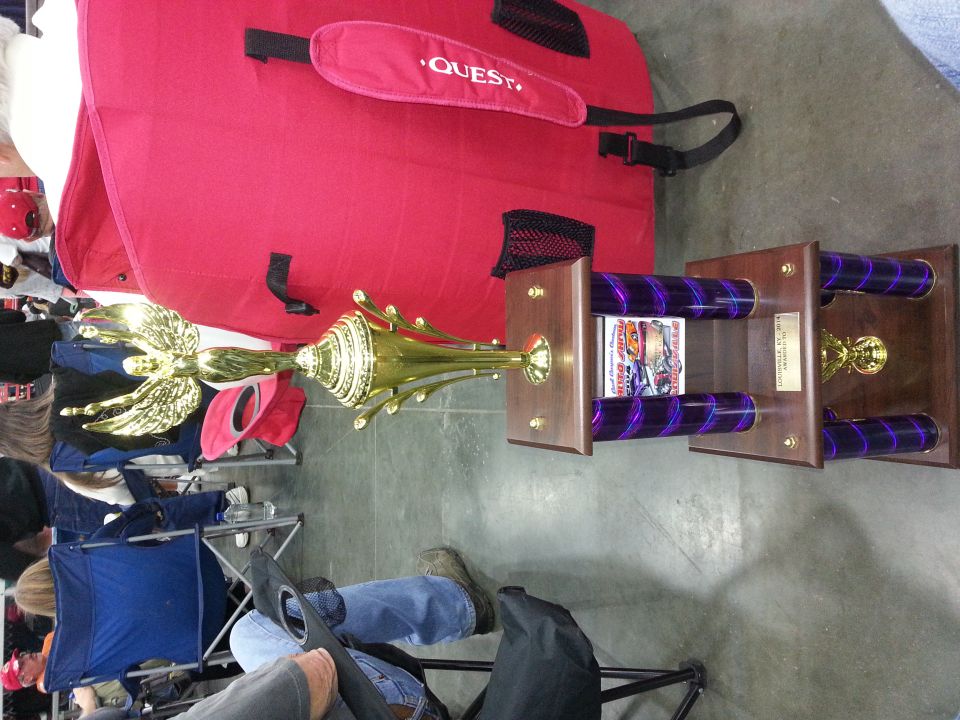 Gold trophy with eagle on top, purple accents, and wooden base. Large red bag with 'QUEST' logo behind it. People sitting in chairs