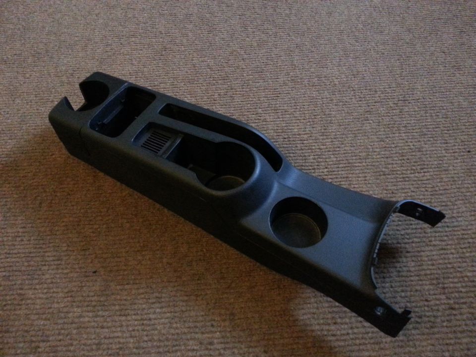 Black plastic center console with two cup holders and storage compartments, lying on a carpeted surface