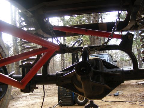 Close-up of a vehicle's rear axle and suspension with red tubing and chains. A welding machine is visible, and