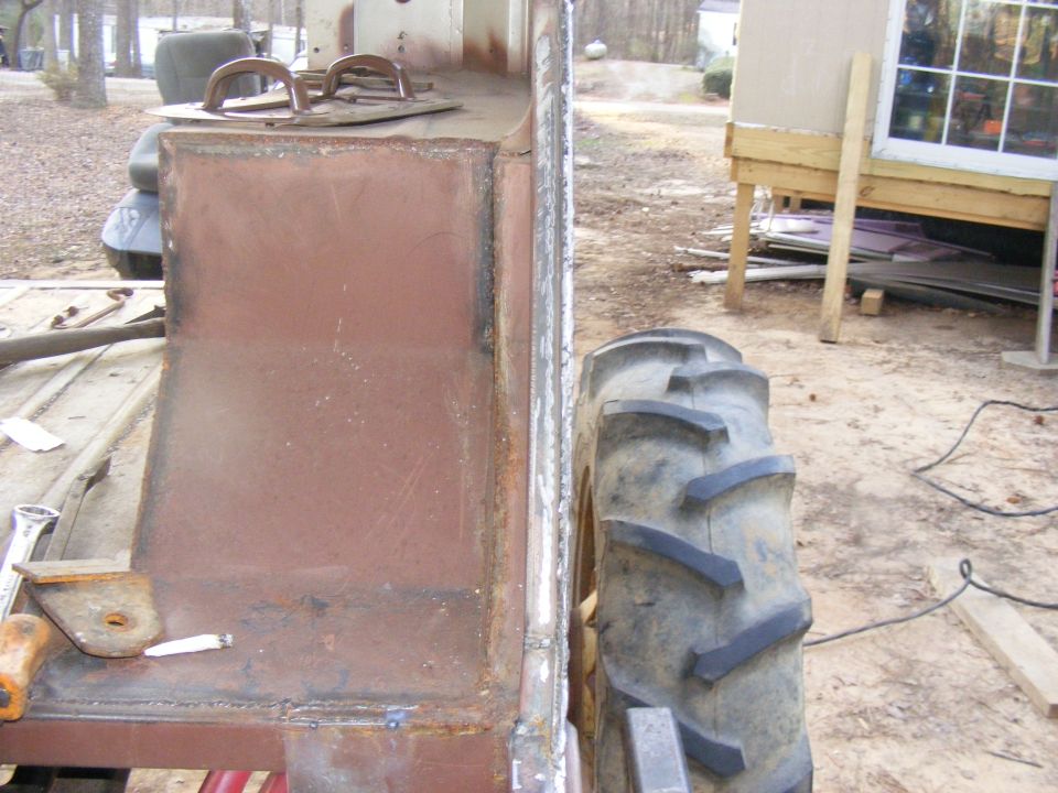 Side view of a custom vehicle build, showing a large tire and unfinished metal body panels. Tools are visible on a wooden surface