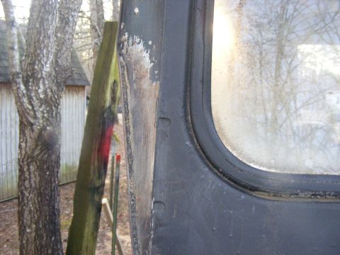 close-up of a dark vehicle door frame with a window, trees and a shed in background