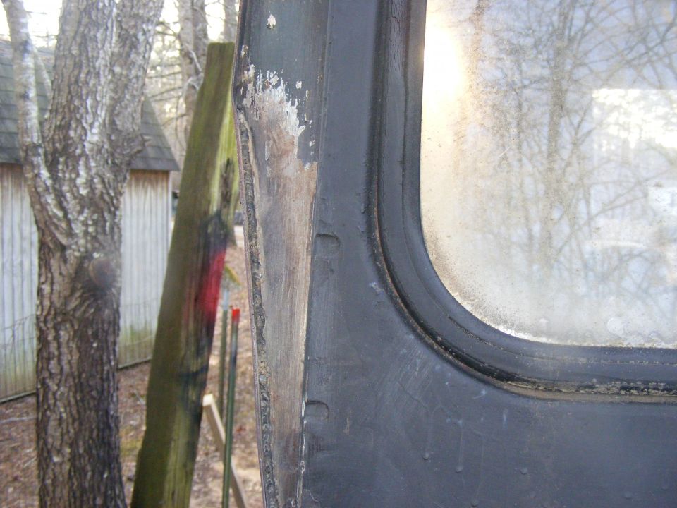 close-up of a dark vehicle door frame with a window, trees and a shed in background