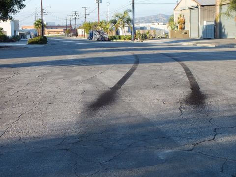 2003 Ford Crown Victoria - Two long curved black tire burnout marks streak across a cracked asphalt street in an industrial area with palm trees and hills behind.