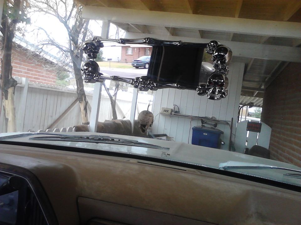 Interior shot of a vehicle looking out the windshield at a custom rearview mirror with skull accents. A car is visible in the background