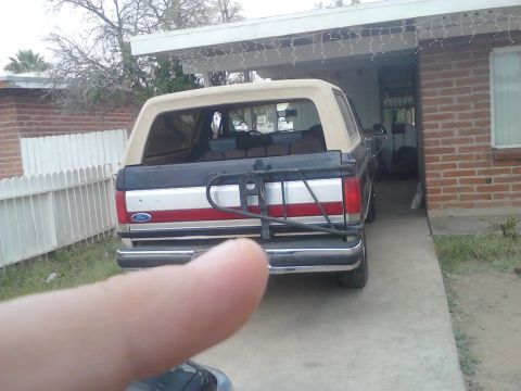 1990 Ford Bronco - The rear of a Ford Bronco is visible, parked under a carport next to a brick house.