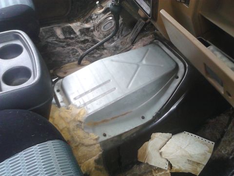 1990 Ford Bronco - The interior floor of a vehicle shows exposed metal and torn insulation, with a cup holder and transmission tunnel cover visible.
