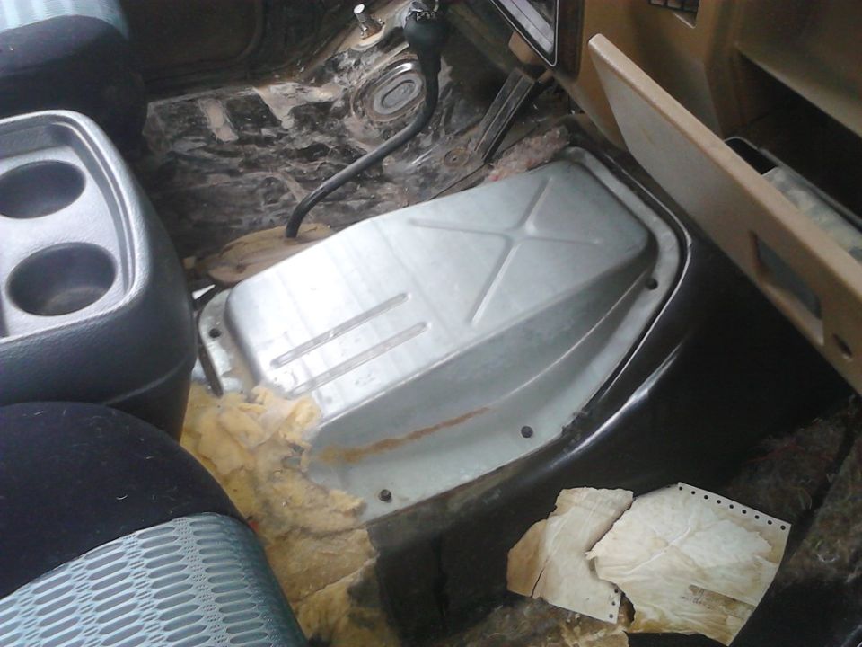 Interior floor of a vehicle with exposed metal and torn insulation, showing a cup holder and part of a transmission tunnel cover