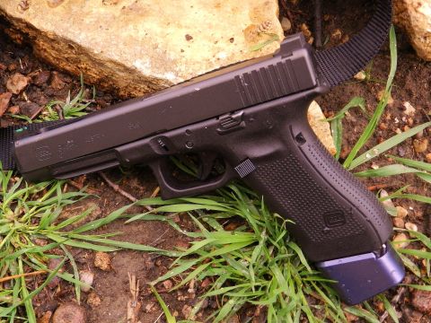 1996 Ford Bronco - A black Glock pistol with a blue extended magazine is shown lying on the ground amongst grass and dirt.
