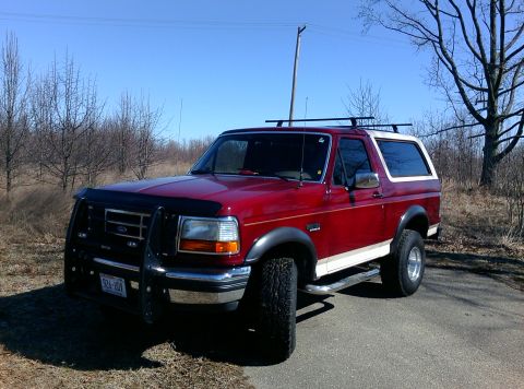 1992 Ford Bronco - A red 1992 Ford Bronco with a white top and a black bull bar is parked on a road with a clear blue sky.