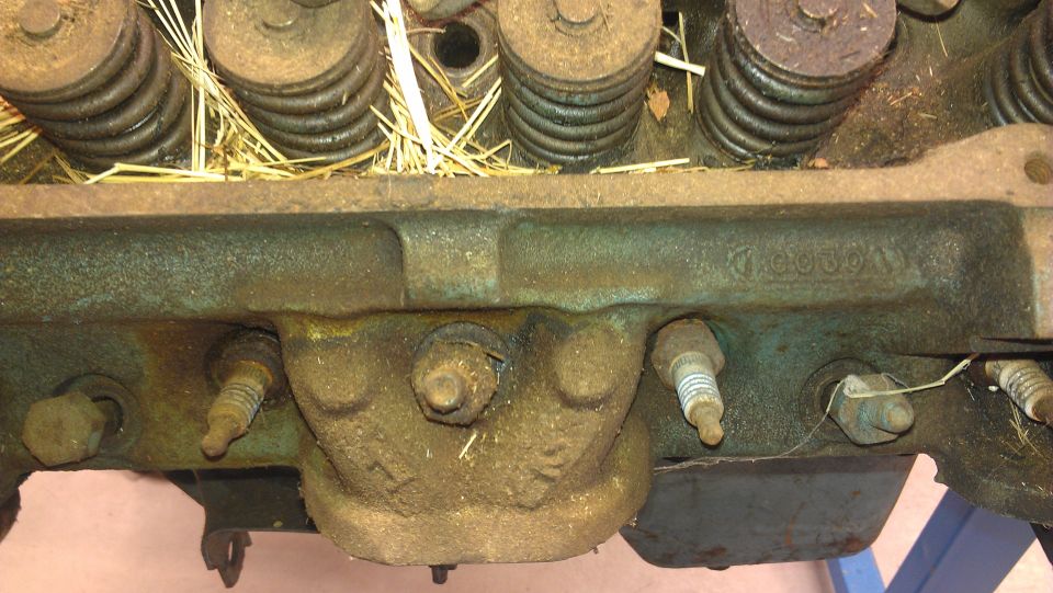 Close-up of an engine cylinder head with spark plugs and valve springs visible. Some straw is present on top