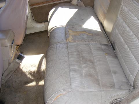 Vehicle interior showing worn, light-colored rear bench seat with torn upholstery and a seatbelt