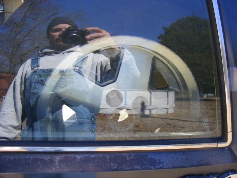 Reflection of man in overalls holding camera, spare tire visible through window