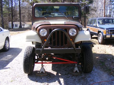 Brown Jeep CJ-5 front view on jack stands, with a wood-paneled SUV behind it