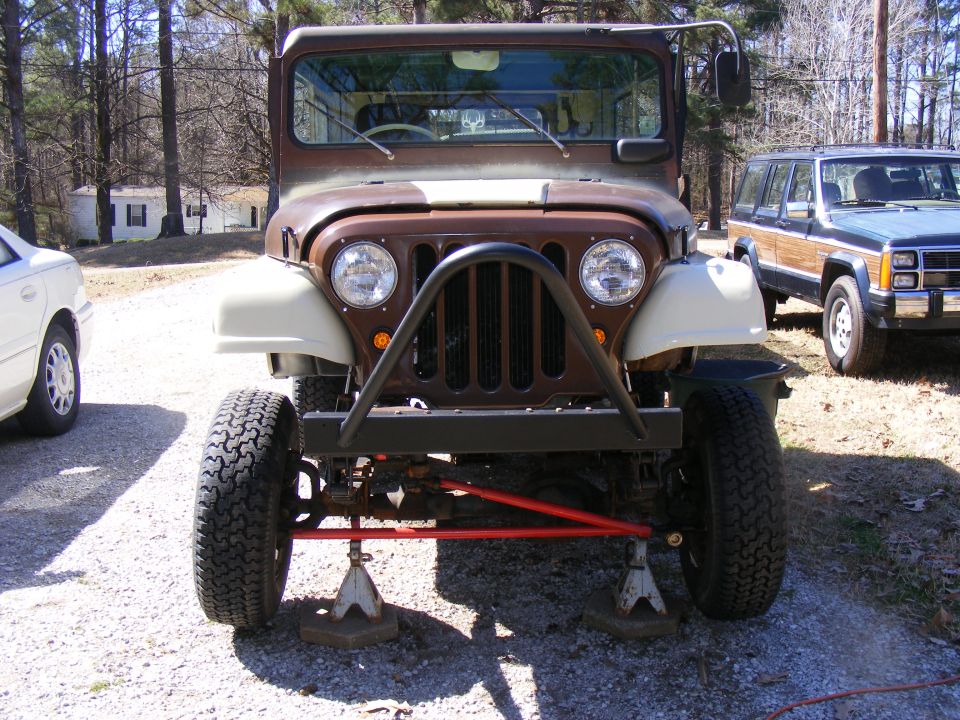 Brown Jeep CJ-5 front view on jack stands, with a wood-paneled SUV behind it