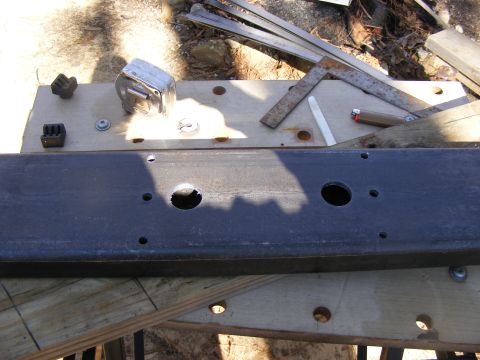 Metal frame part with two large circular holes and several smaller ones, resting on a wooden surface with tool