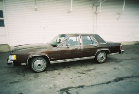 1984 Mercury Grand Marquis - A brown sedan is parked on a wet concrete surface in front of a white building.