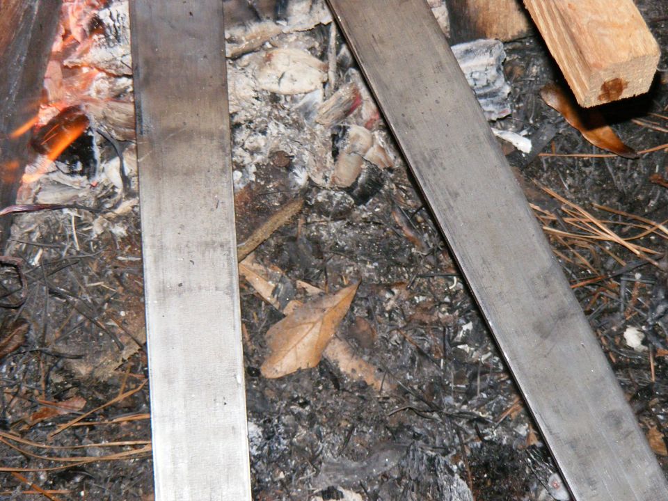 Two metal bars lie across campfire ashes with burning embers visible on the left
