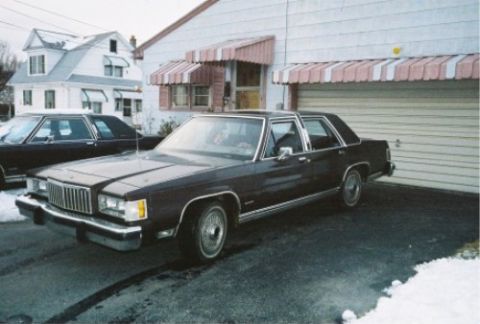 1984 Mercury Grand Marquis - A dark sedan with a prominent chrome grille is parked in a driveway next to another dark sedan.