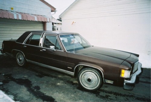 Brown four-door full-size sedan with vinyl roof and wire wheel covers parked by a white garage