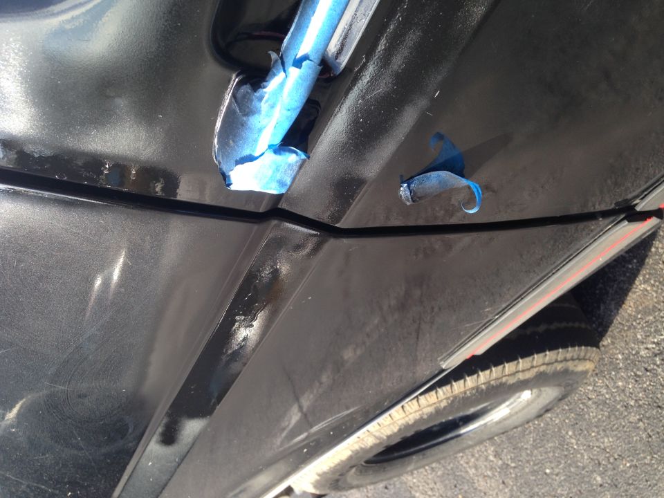 Close-up of a black car's door seam with blue painter's tape. A tire is visible at the bottom