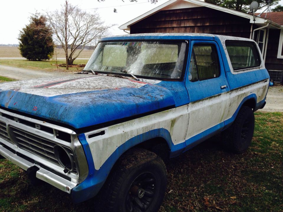 Blue and white Ford Bronco with peeling paint and missing headlight, parked on grass