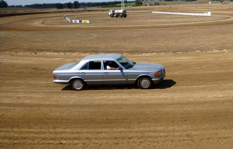 Silver sedan driving on a dirt track with a water truck in the background