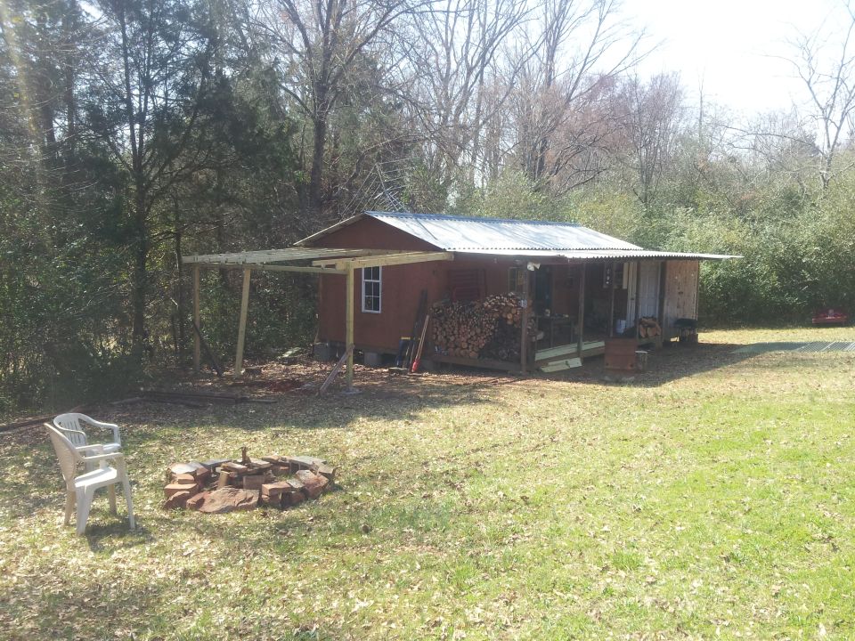 Cabin with a covered porch and stacked firewood, next to a fire pit and a white plastic chair