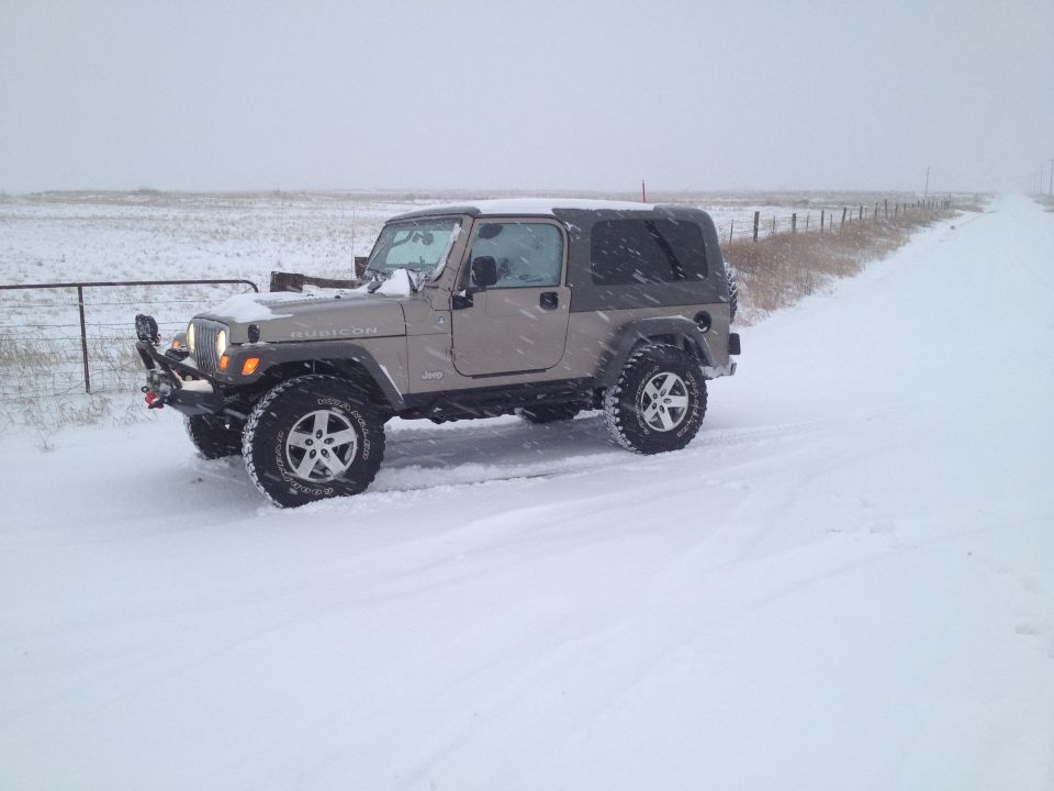 Tan Jeep Wrangler with a black soft top driving on a snowy road next to a fence