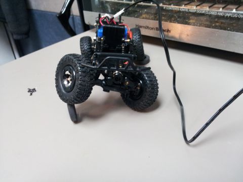 1995 Ford Bronco - A black remote control car chassis with exposed wiring and large tires sits on a table next to a power cord.