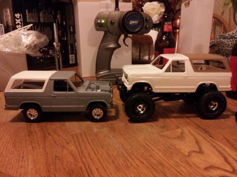 1995 Ford Bronco - Two model RC cars, a gray and a white Bronco, sit on a wooden surface with a remote control nearby.