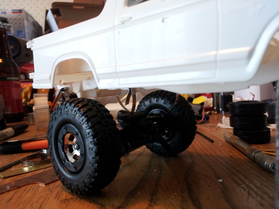 White model car body on a chassis with large tires, sitting on a wooden surface with tools
