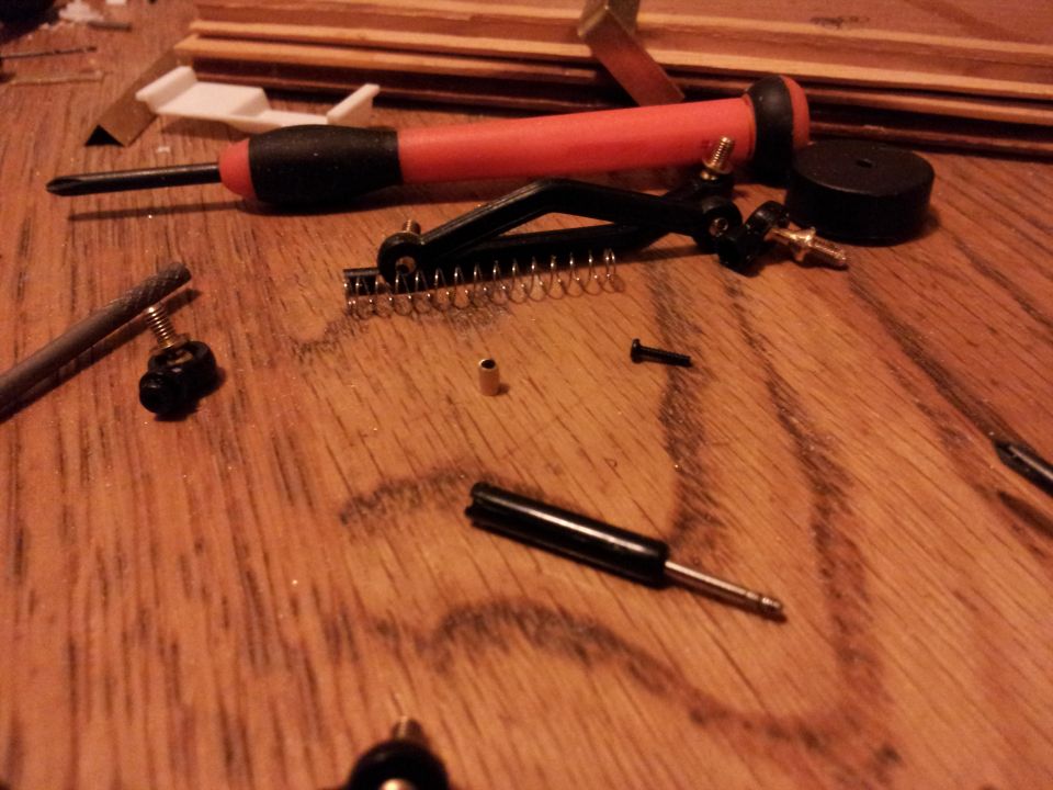 Model car parts and tools including a screwdriver, springs, and screws scattered on a wooden surface