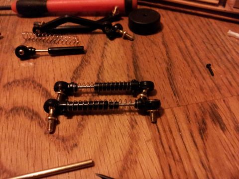 1995 Ford Bronco - Two assembled suspension linkages, likely for a model car, lie on a wooden surface alongside tools and springs.