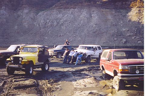 1992 Ford Bronco - Several off-road vehicles are stuck in the mud, with people attempting to push them free.